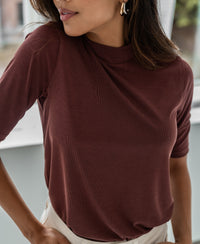 Woman wearing a ribbed collared top in cacao color, by Plein Publique, standing indoors near a window.