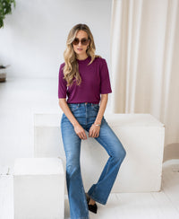 Woman wearing light blue flared jeans sits on a white block in a bright room.