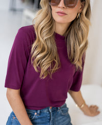 Woman wears a magenta ribbed collared top indoors.