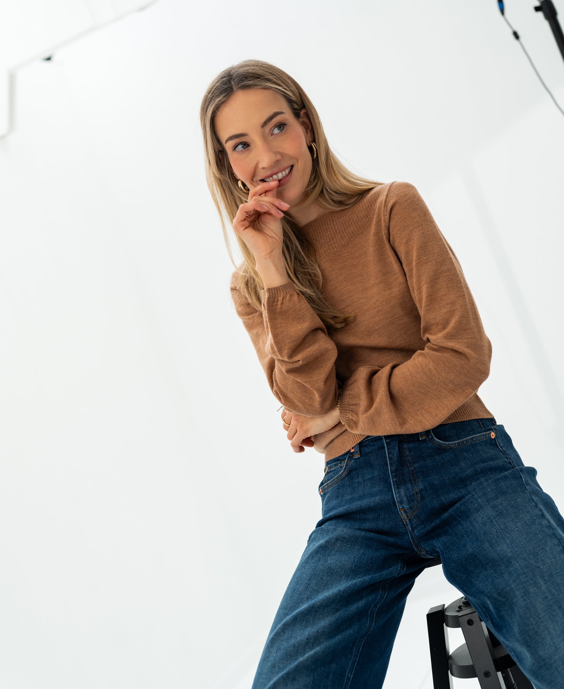 Woman wearing PLEIN PUBLIQUE's LA MUSE Chai tea boat neck sweater leans on a stool in a bright studio.