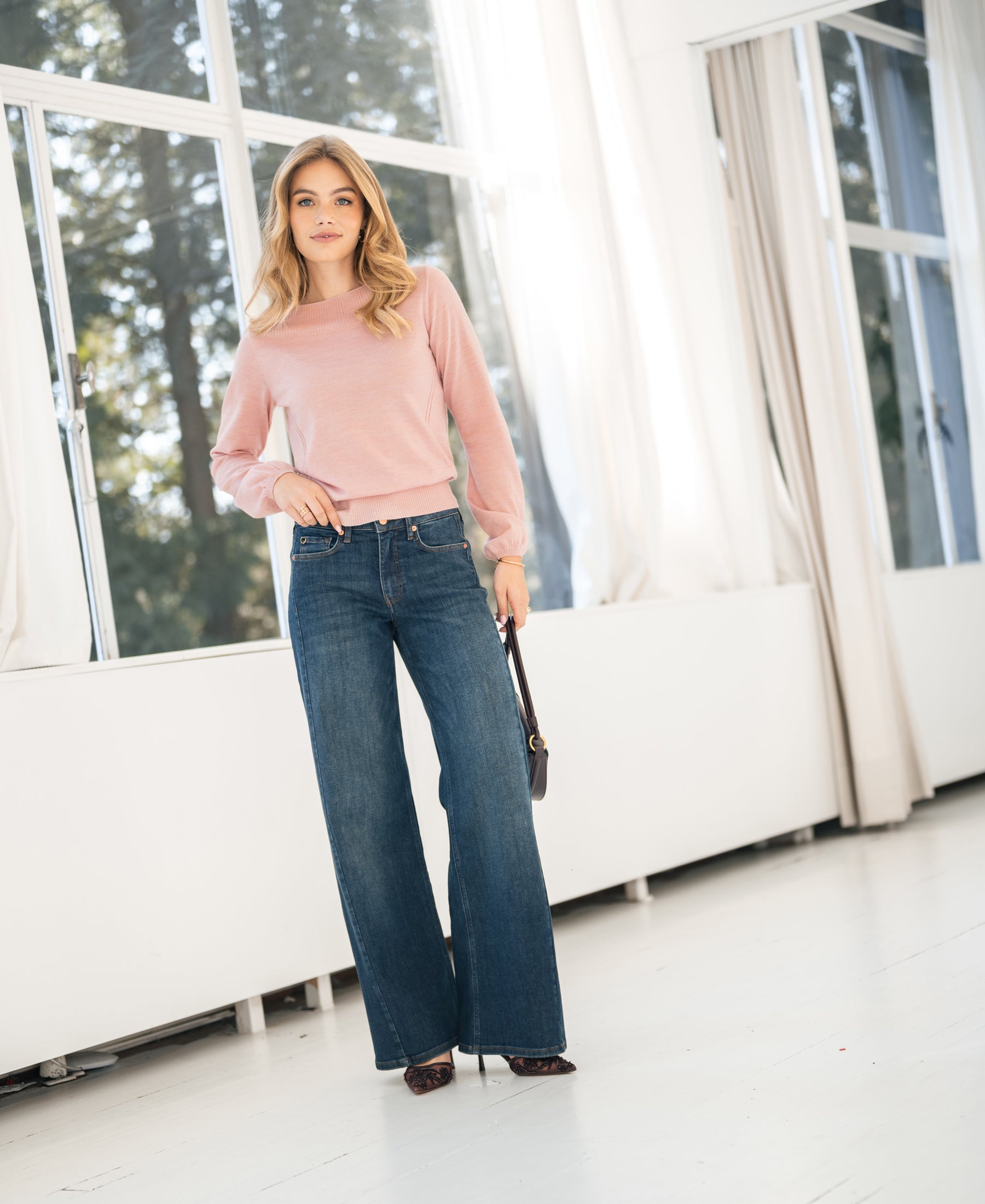 Woman indoors by large windows wearing light pink boat neck sweater.