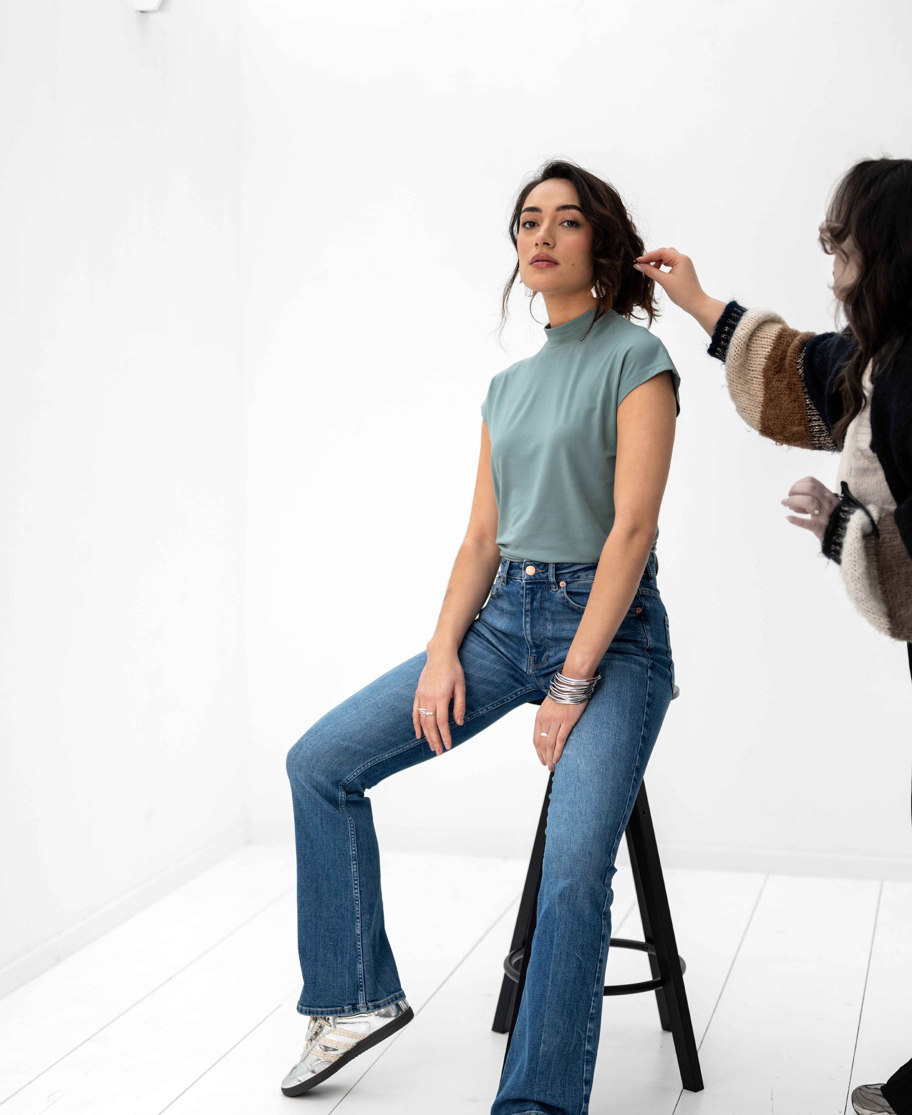 Woman wearing the PLEIN PUBLIQUE LA ROMY Travel Top in light green sits on a stool in a bright room.