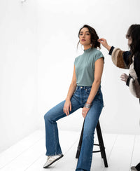 Woman wearing the PLEIN PUBLIQUE LA ROMY Travel Top in light green sits on a stool in a bright room.