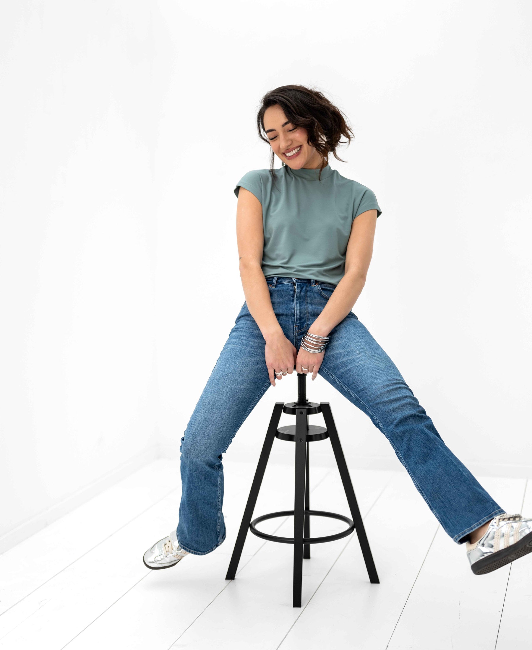 Woman wearing light green PLEIN PUBLIQUE LA ROMY travel top sits on a stool, smiling with legs playfully extended.