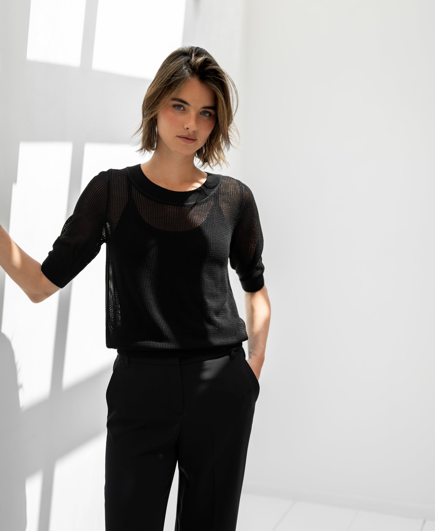 Woman wears Plein Publique LE NANCY mesh silk sweater in black, posing by a sunlit white wall.