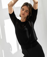 Black mesh sweater with silk by PLEIN PUBLIQUE shown on a woman, eyes closed, sunlight on the wall behind.