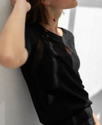 Woman wearing black mesh sweater LE NANCY with silk by PLEIN PUBLIQUE, leaning on a white wall in bright light.