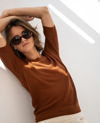 Woman wearing a mesh sweater with silk leans against a white wall, sunlight casting shadows.