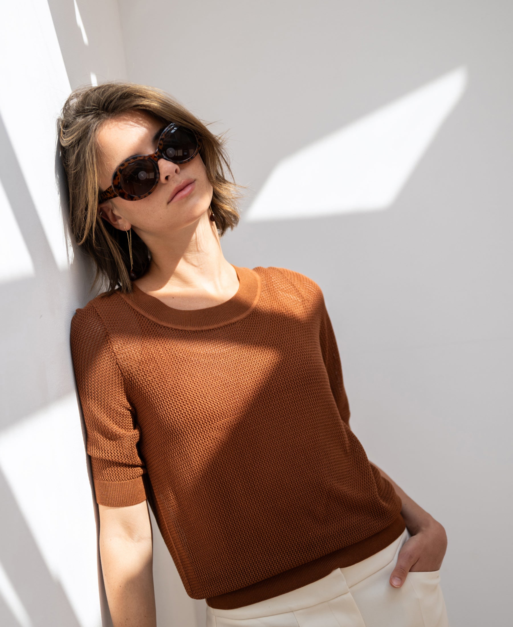 Mesh sweater LE NANCY with silk in Havana by PLEIN PUBLIQUE, worn by a woman leaning against a sunlit wall.