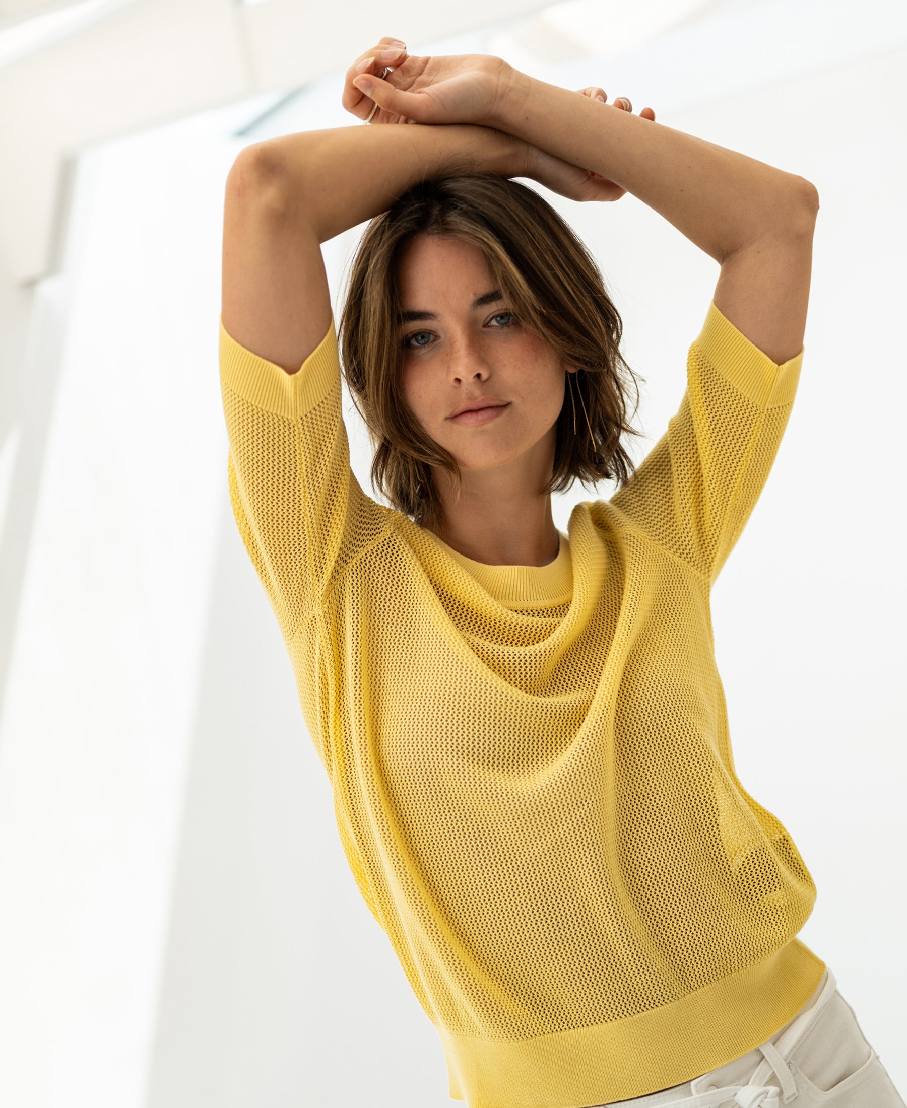 Woman wears lemon yellow PLEIN PUBLIQUE mesh sweater with silk, arms raised indoors.