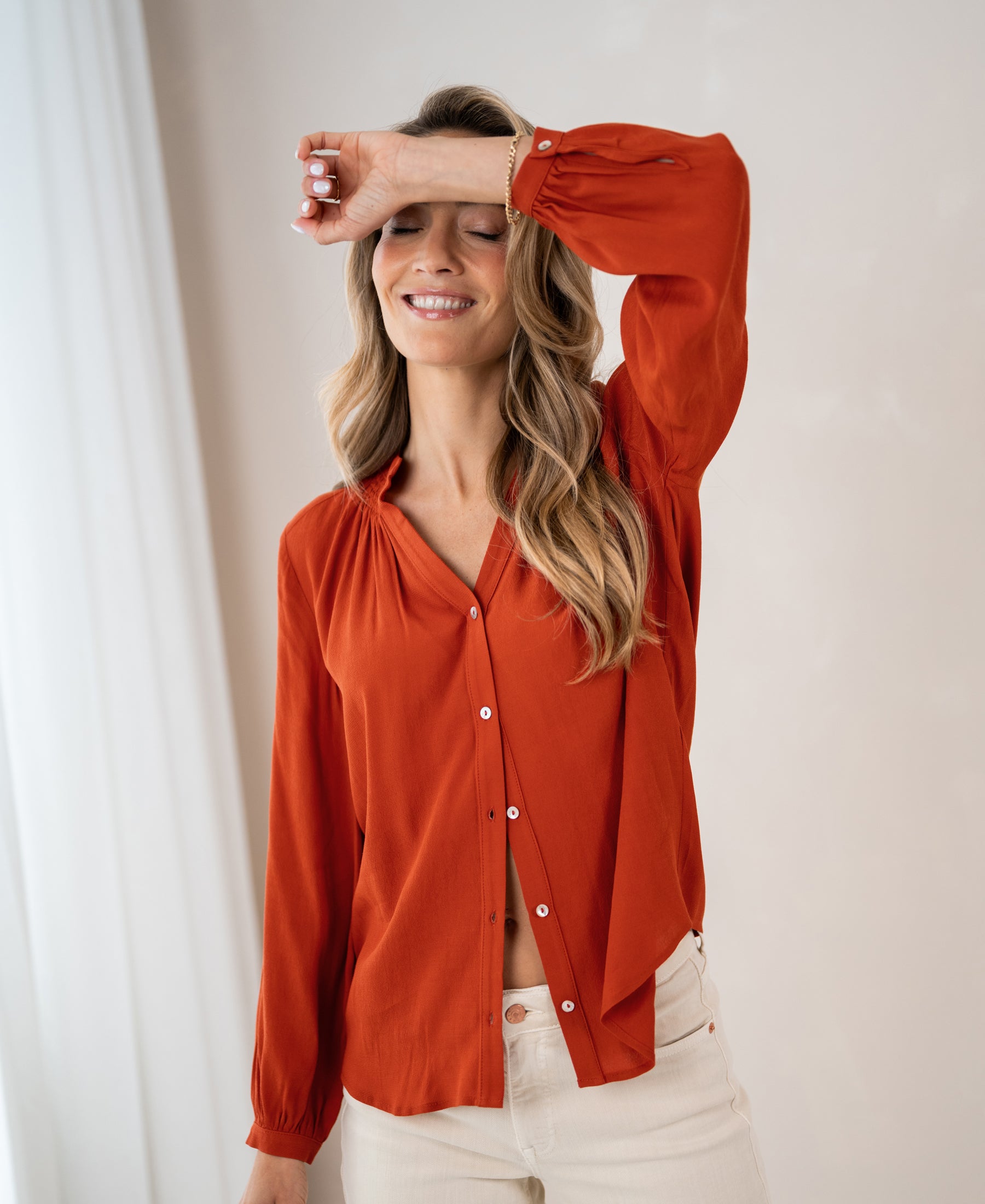 Woman wearing LA NIMES burnt orange blouse by PLEIN PUBLIQUE, standing with hand on forehead in bright room.