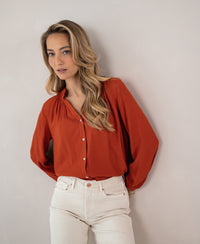 Burnt orange LA NIMES blouse by PLEIN PUBLIQUE shown against a light wall.