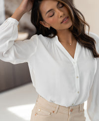 Woman wearing a white LA NIMES blouse indoors, eyes closed, gently touching her hair.