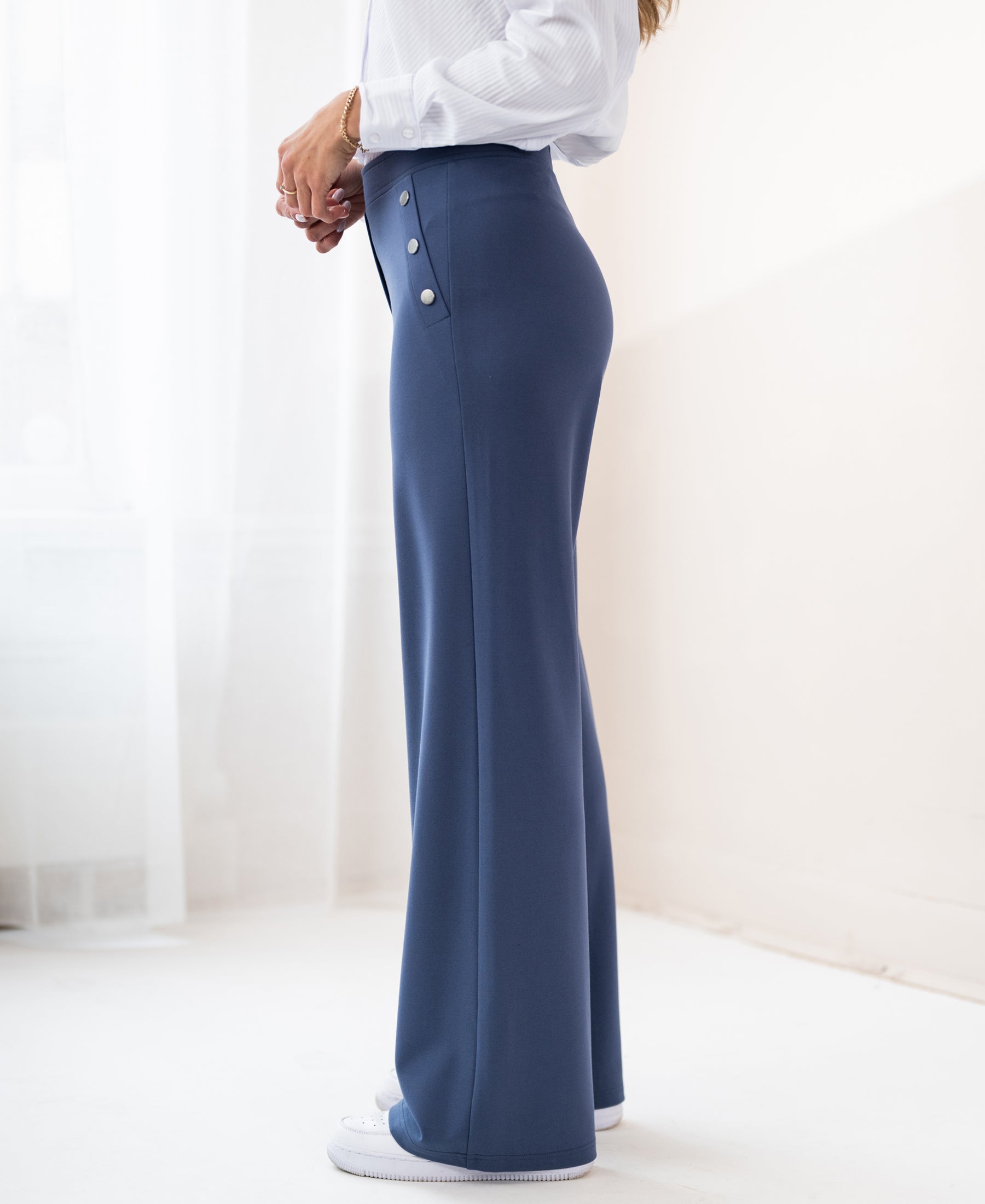 Woman wearing blue PLEIN PUBLIQUE Travel Pants LA NOAN Deluxe, standing in a bright room.
