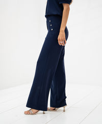 Woman wears navy blue PLEIN PUBLIQUE travel pants called LA NOAN Deluxe on a white floor.