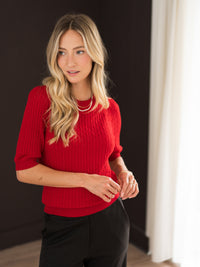 Red LA NONNA short-sleeve cable sweater displayed on a woman standing by white curtains.