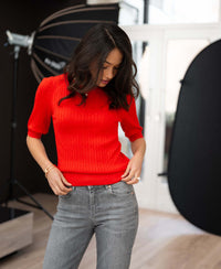 Woman wears coral-red cable knit short-sleeve sweater, adjusts it while standing in studio.