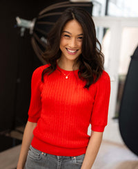 Coral red short-sleeve cable knit sweater in a bright, sunlit room.