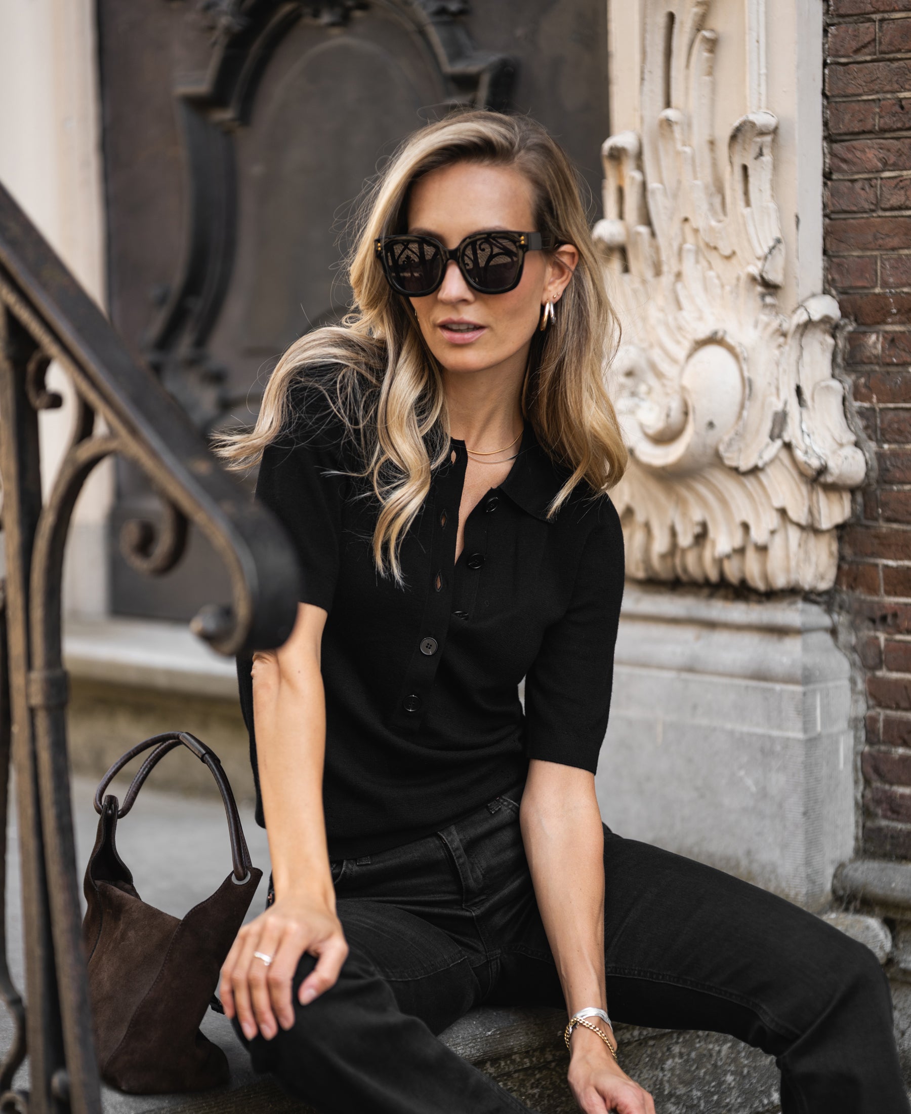 Woman wearing a black merino wool polo sweater by PLEIN PUBLIQUE sits on steps.