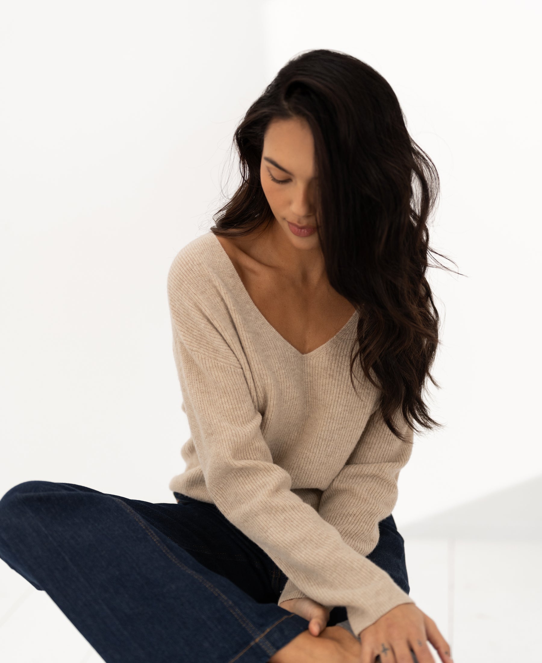 Relaxed merino wool sweater in light sand color, shown on model with head bowed, in a bright room.