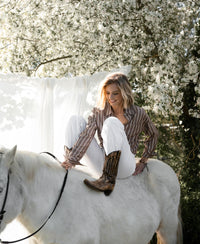 Woman wearing a PLEIN PUBLIQUE Tencel blouse in brown-ivory stripes sits side-saddle on a white horse.