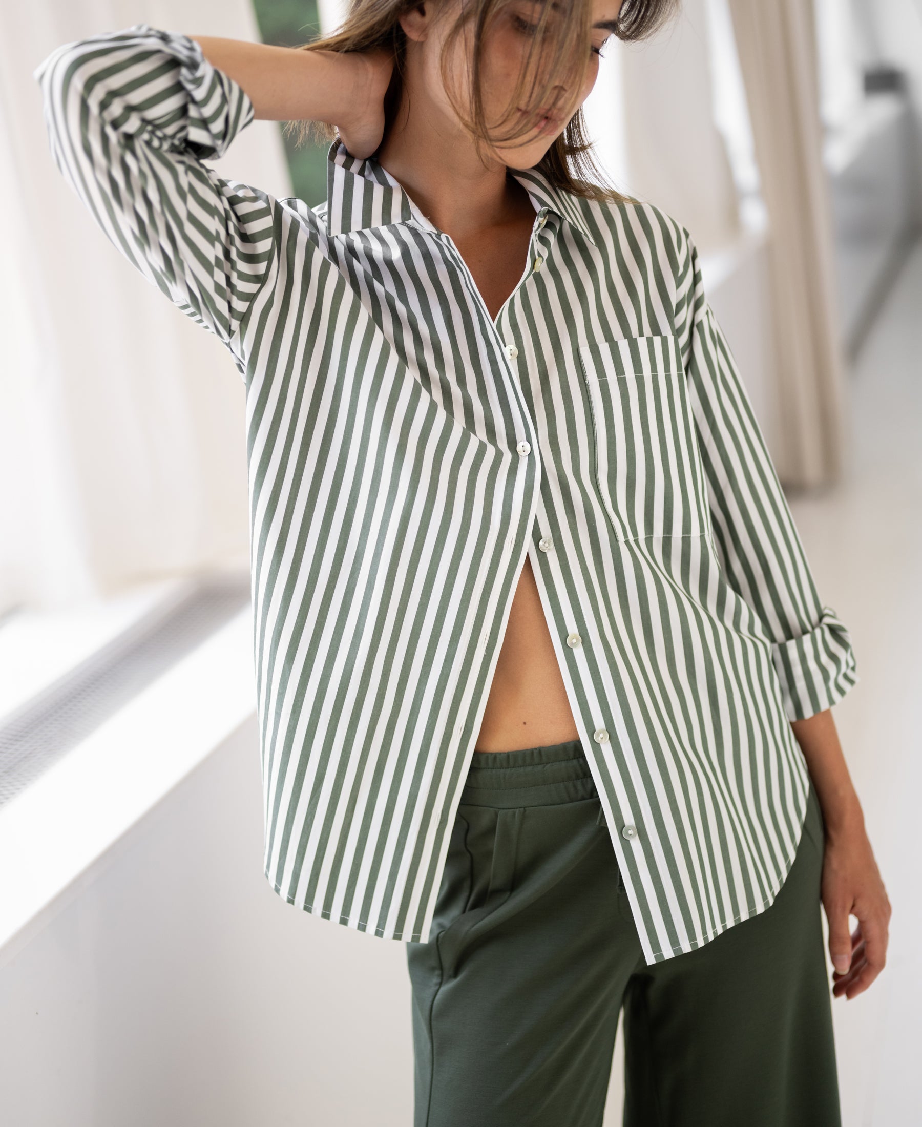 Woman wearing a PLEIN PUBLIQUE LA PASCALLE white and olive-green striped blouse indoors by a sunlit window.