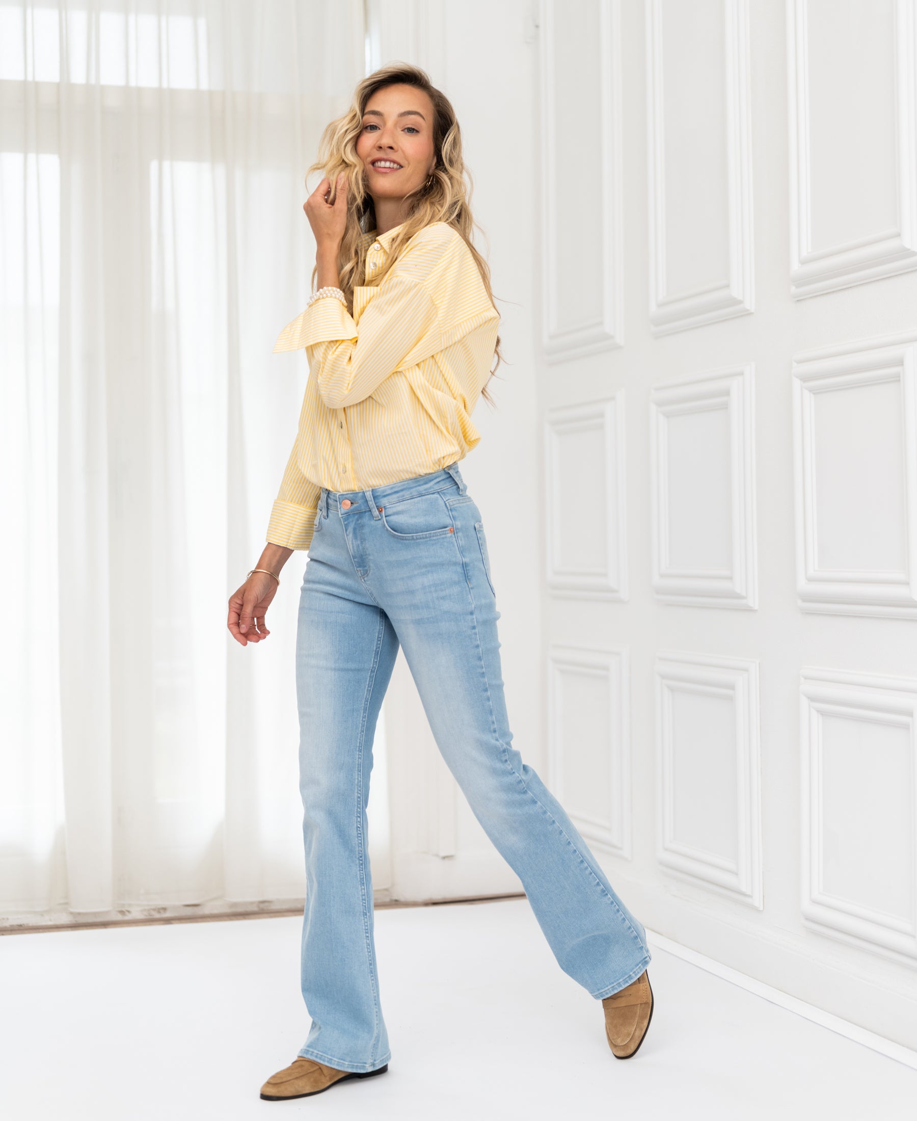 Woman wears OVERIG’s Les Flares bleached jeans, posing in a sunny room with large windows.