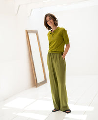 Olive green Merino wool-silk polo shirt shown on a woman in a bright, minimalist room with a floor mirror.
