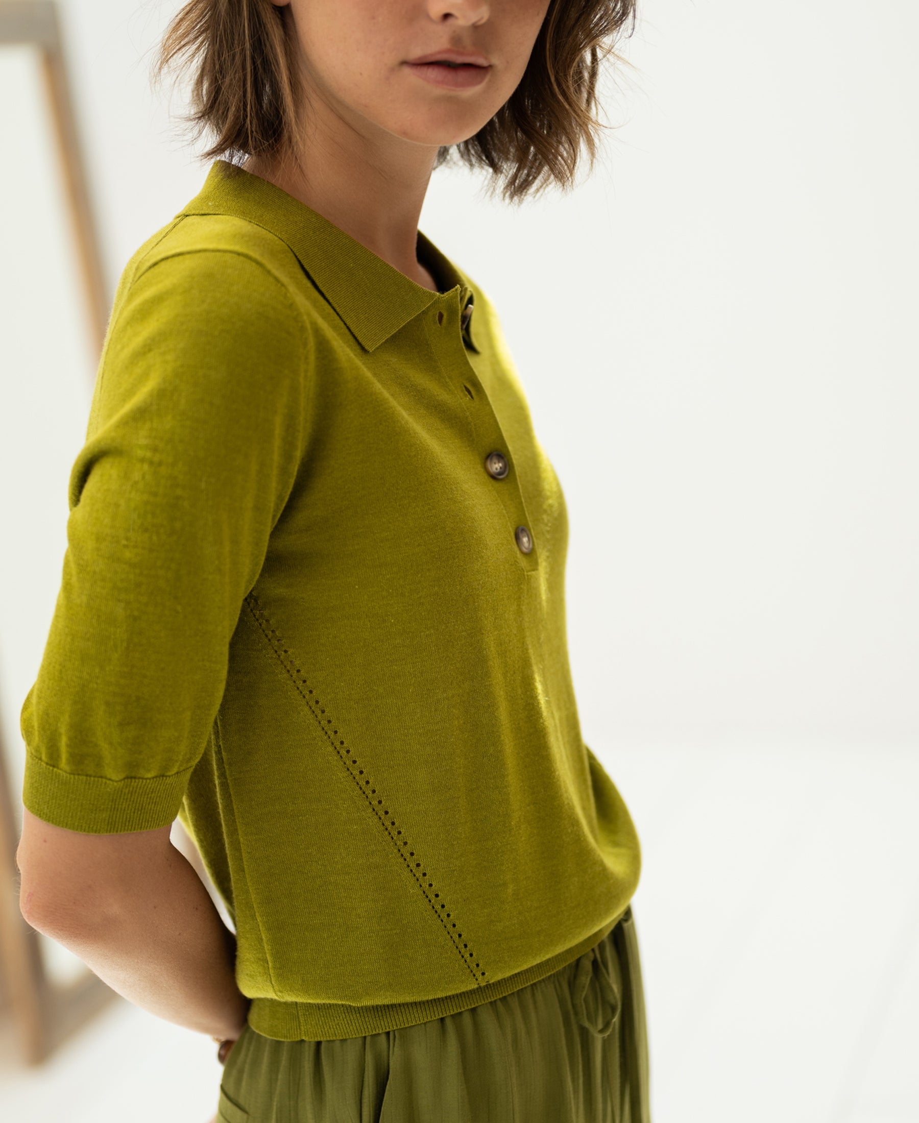 Woman wears the Merino wool-silk polo shirt in olive green by PLEIN PUBLIQUE, shown in soft lighting.