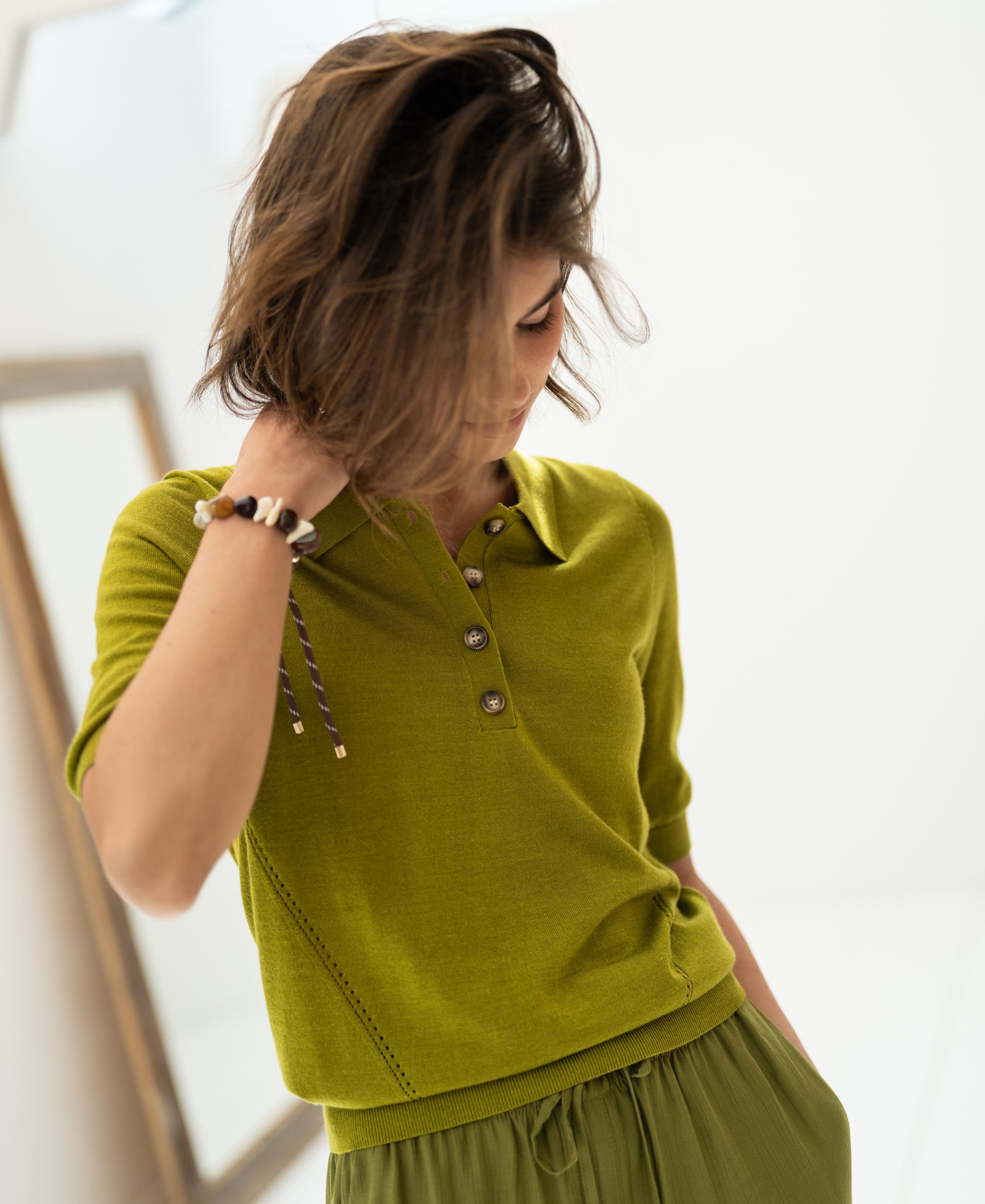 Woman wearing an olive green merino-silk polo shirt looks down, touching her hair in a sunlit room.