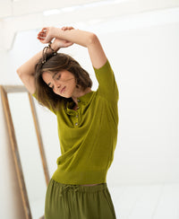 Woman wearing an olive green Merino-silk polo shirt stands with arms raised, eyes closed, next to a mirror.