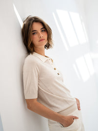 Woman wearing a PLEIN PUBLIQUE merino wool-silk polo shirt in sand-ivory leans on a white wall.