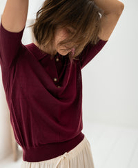 Woman wearing the PLEIN PUBLIQUE merino wool-silk polo shirt in wine, arms raised, hair partly covering her face.