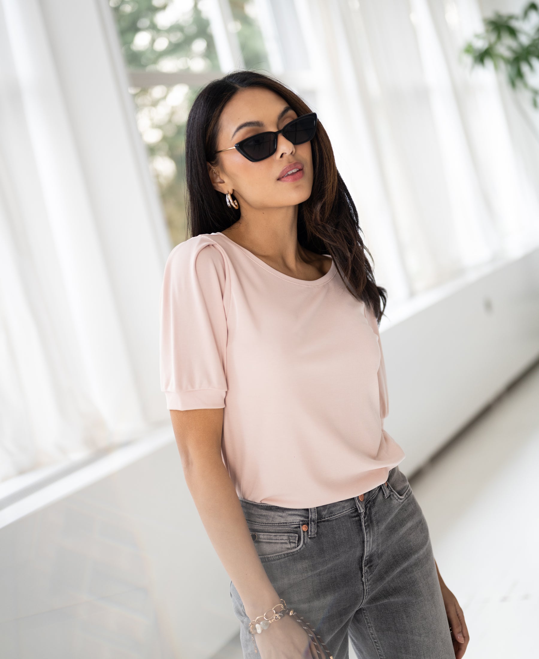 Woman wearing a ribbed blush PLEIN PUBLIQUE LA PHILEINE T-shirt stands in a bright room with white curtains.