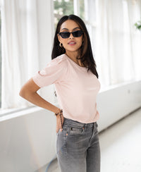 Woman wearing a ribbed blush T-shirt stands indoors by large windows with white curtains.