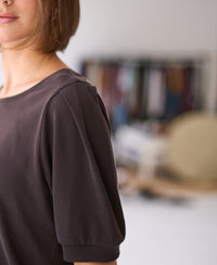 Woman wearing a ribbed chocolate brown T-shirt indoors, with colorful fabrics softly blurred in the background.