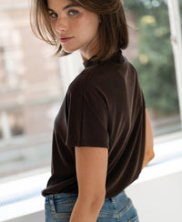 Woman wearing a chocolate brown ribbed top looks over her shoulder by a bright window.