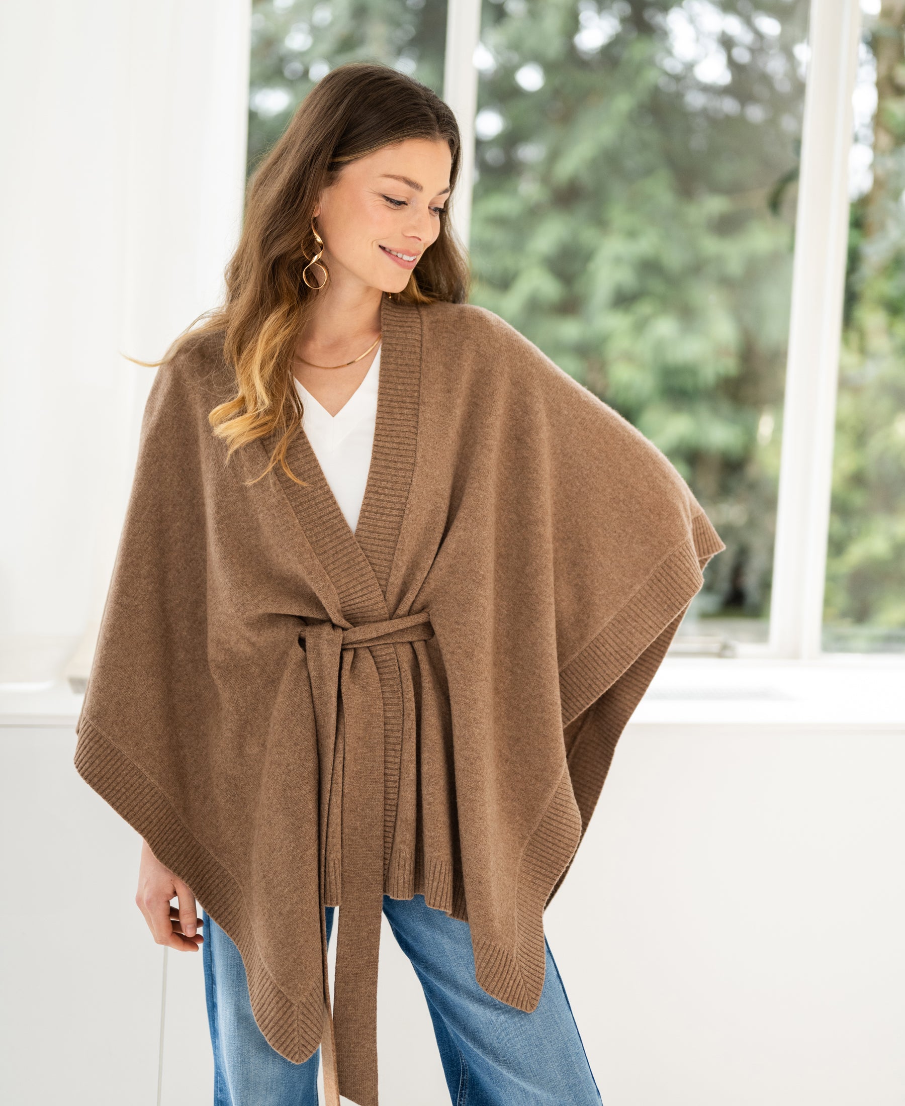 Camel merino wool poncho in a relaxed fit, shown on a person standing by a window with greenery outside.