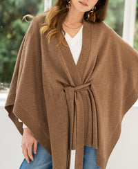 Camel merino wool poncho by PLEIN PUBLIQUE, shown on a woman standing by windows.