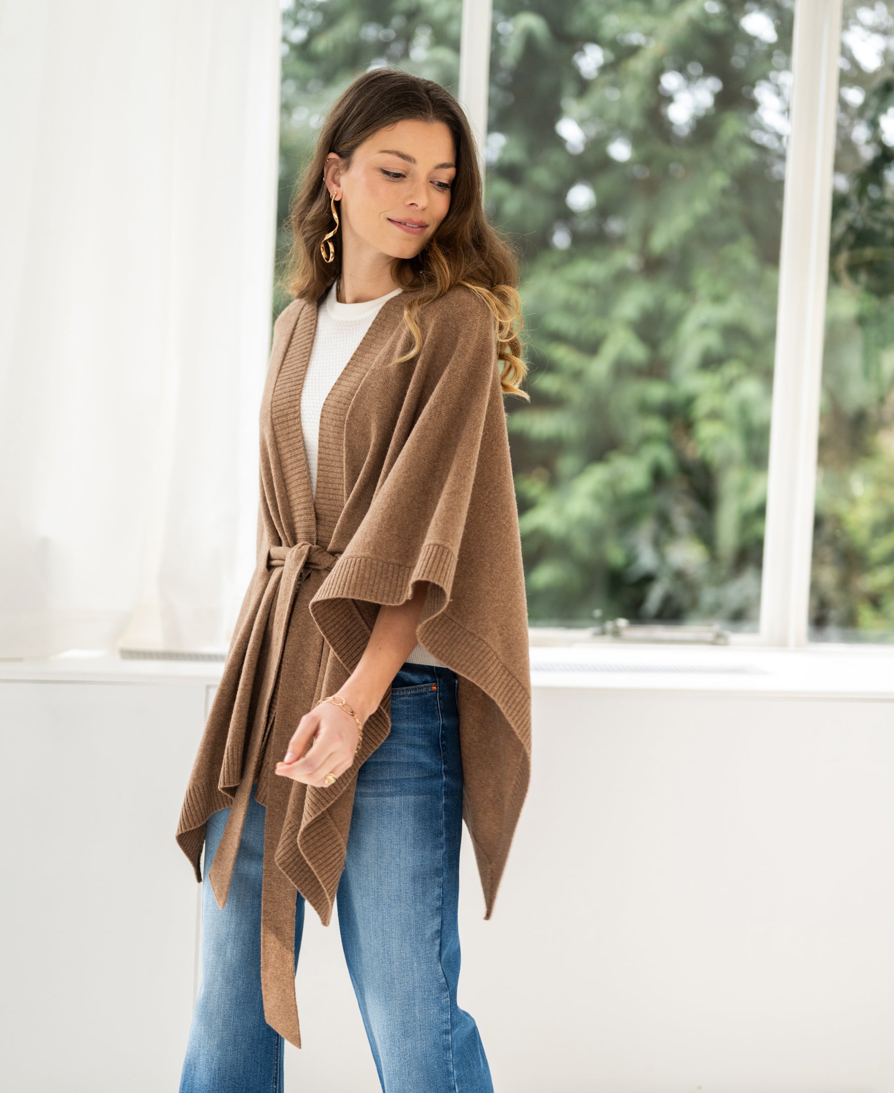 Camel merino wool poncho by PLEIN PUBLIQUE, shown on a woman standing by a window with greenery outside.