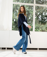 A dark blue merino wool poncho by Plein Publique displayed near sunlit windows with greenery in the background.