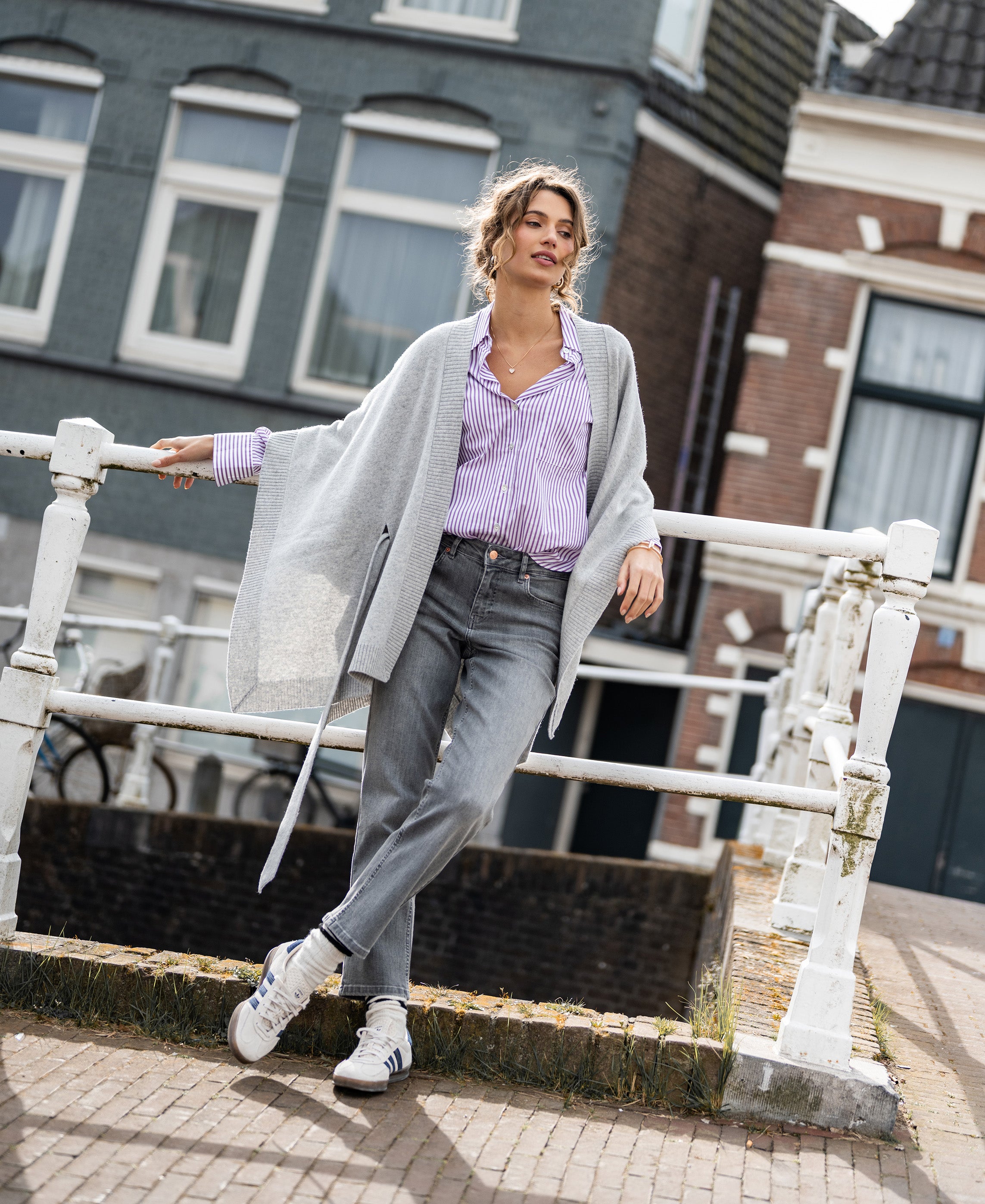 Merino wool poncho in silver grey by PLEIN PUBLIQUE, shown worn outdoors while leaning on a white railing.