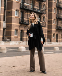 Black merino wool poncho by PLEIN PUBLIQUE shown on a woman outdoors, with brick buildings in the background.