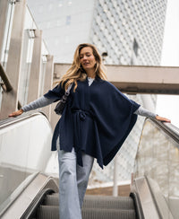 Dark blue PLEIN PUBLIQUE merino wool poncho shown outdoors on a descending city escalator.