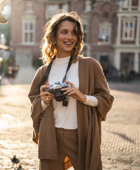 Woman wearing a camel-colored merino wool poncho stands outdoors, smiling and holding a camera.