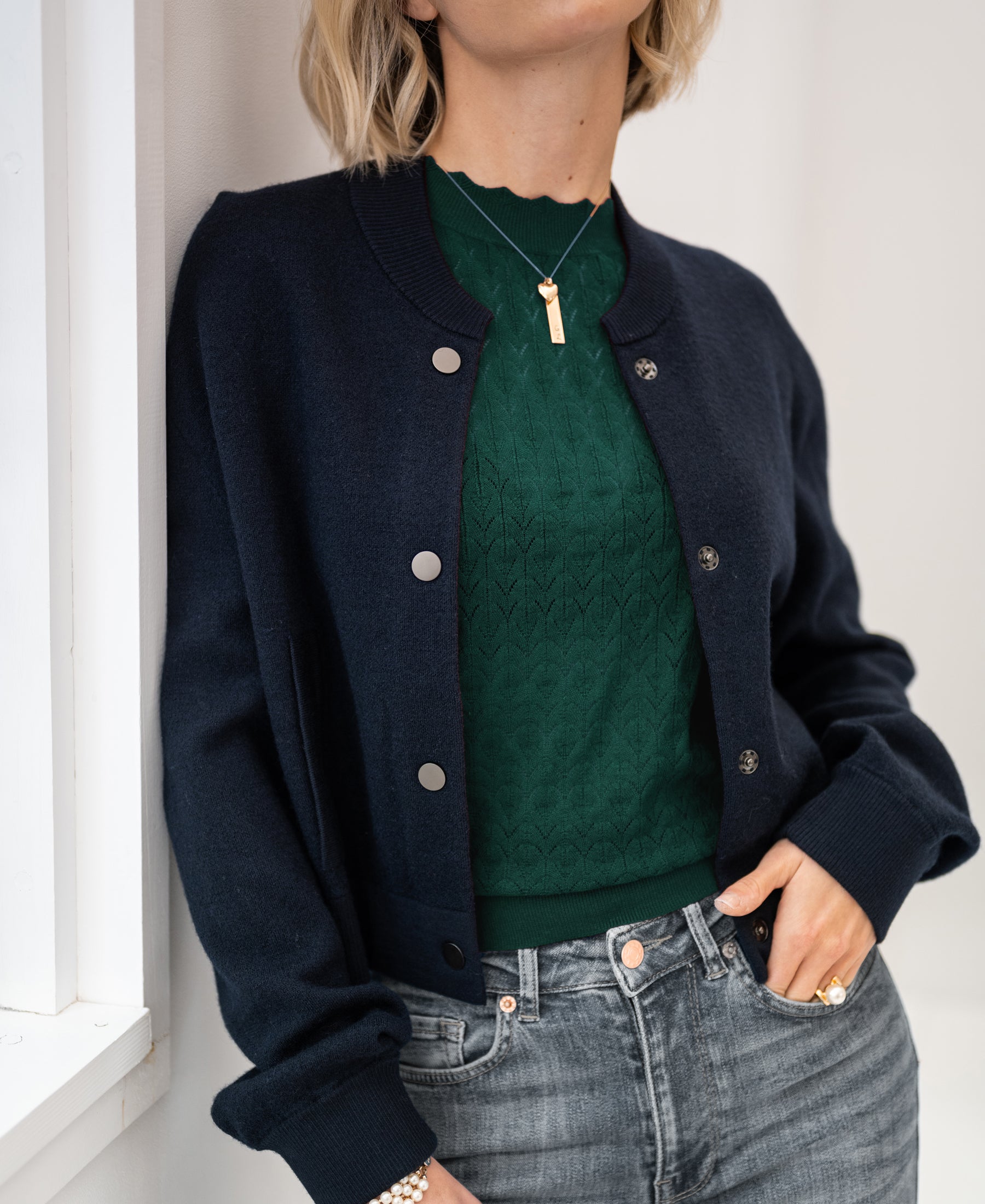 Woman wearing a dark blue LA BOMBA bomber jacket, standing by a white wall.