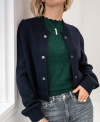 Woman wearing a dark blue LA BOMBA bomber jacket, standing by a white wall.