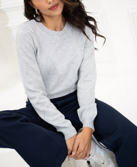 Woman wearing a Merino blend sweater in light grey by PLEIN PUBLIQUE, sitting with hands on knees.