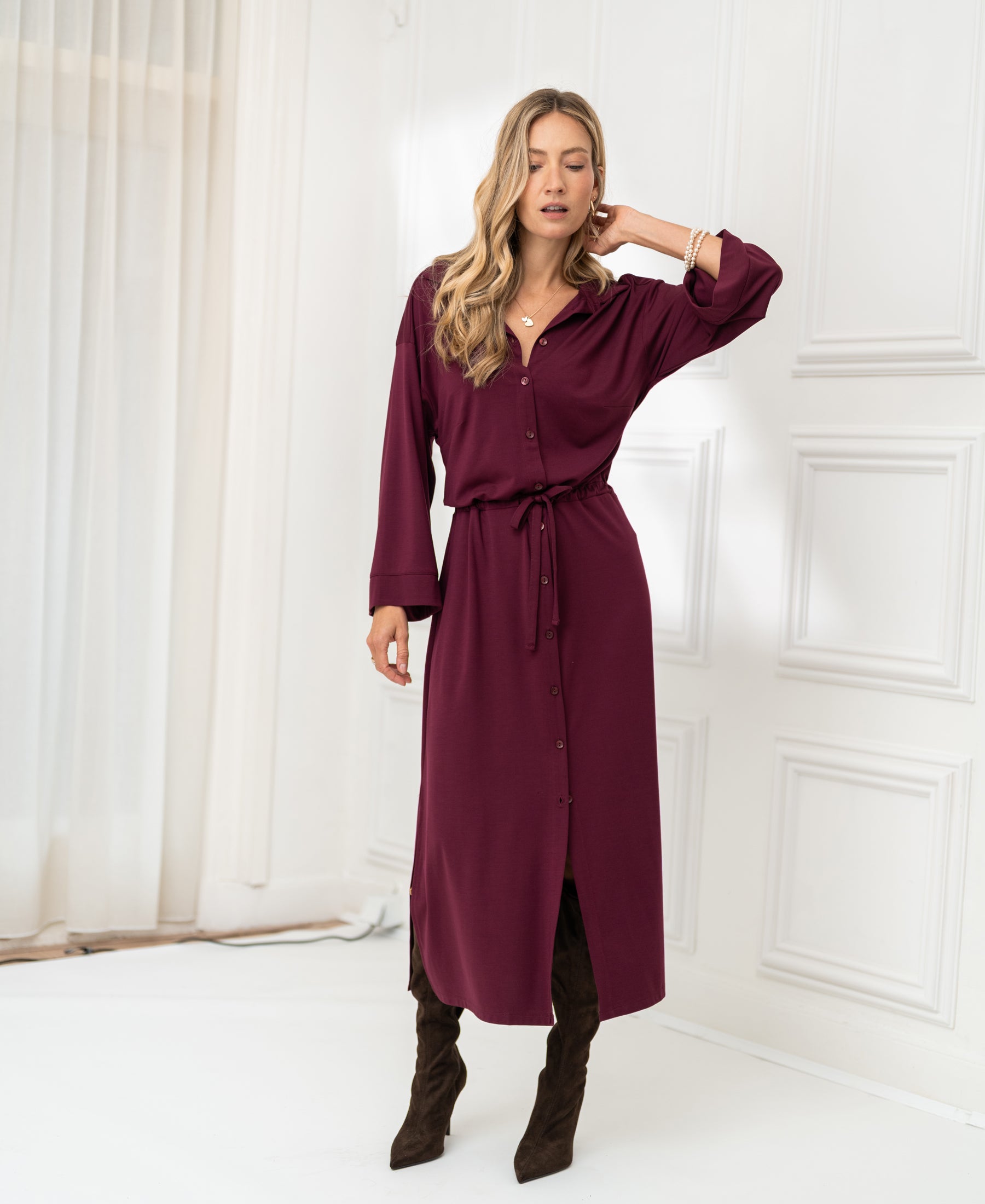Woman wearing a Bordeaux LE RAPHAEL button-front dress with tie waist by PLEIN PUBLIQUE, standing indoors.