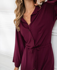 Front view of a woman wearing a Bordeaux button-down dress by PLEIN PUBLIQUE, standing against a white wall.